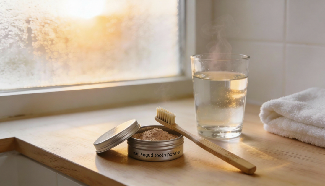 A calm morning bathroom counter with an open metal tin labeled “nanjangud tooth powder,” a bamboo toothbrush, a glass of warm water, and a folded white towel in soft sunrise light.